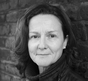 Black and white portrait of Emily Mayhew. Emily is wearing a black jacket and stands smiling in front of a brick wall