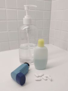 A bottle of hand sanitizer, another of shampoo, an asthma inhaler and a handful of white tablets on a bathroom shelf.